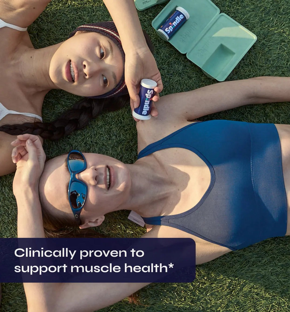 Two women lying on grass with a Spindle Muscle+ bottle, clinically proven to support muscle health.