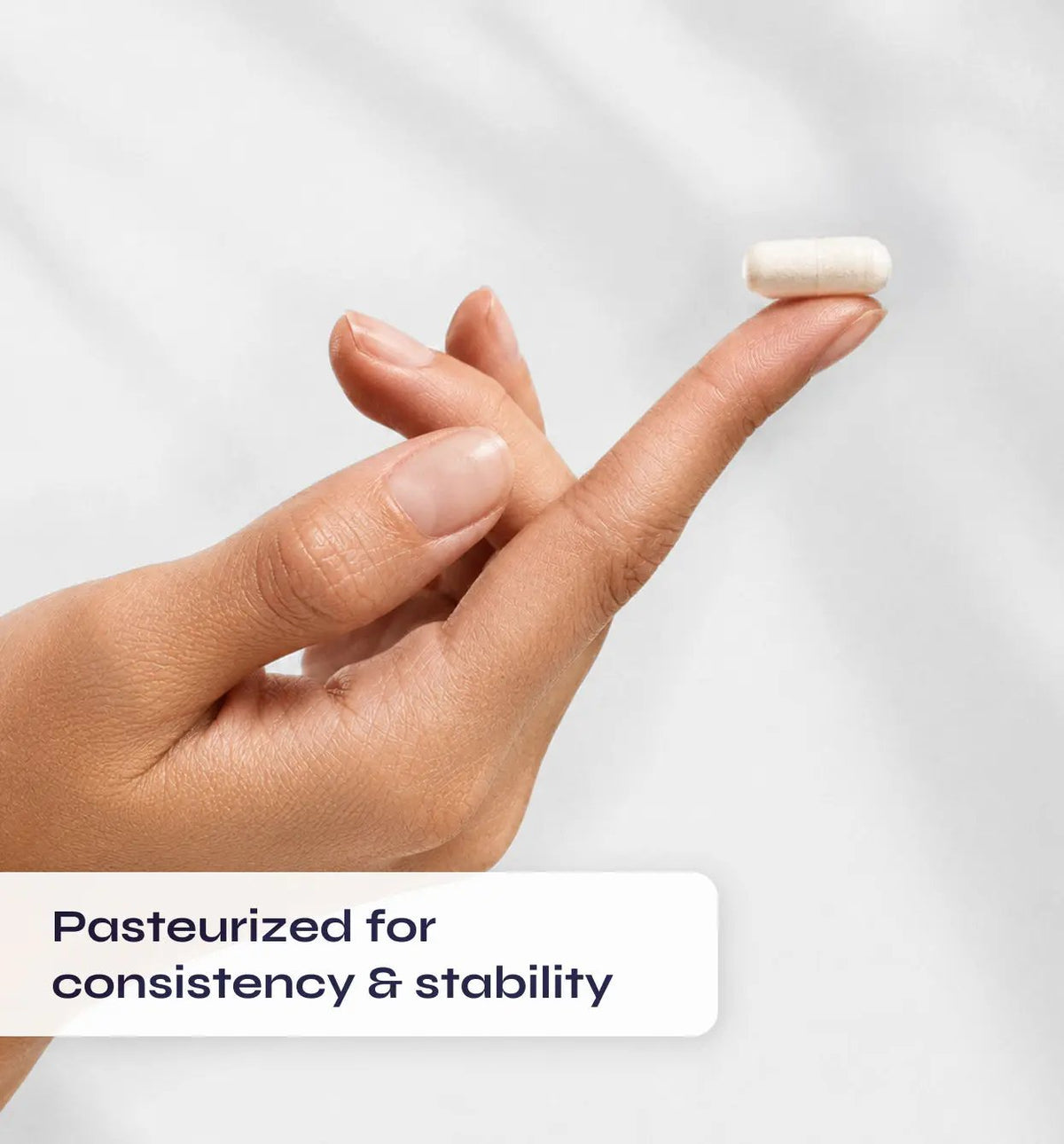 Hand holding a small white pill with text 'Pasteurized for consistency & stability' on a light gray background