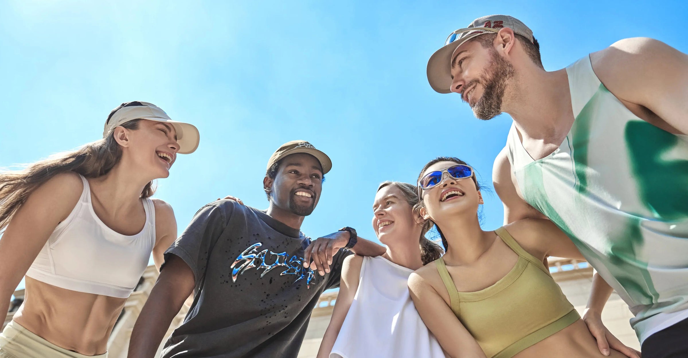 Five diverse friends wearing athletic wear and caps laughing together in a circle under a bright blue sky.